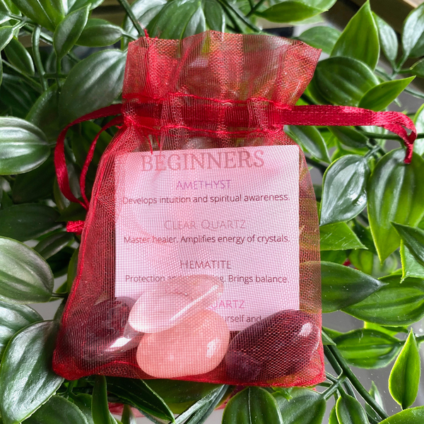 A selection of four crystals inside a red organza bag, crystals include Amethyst, Clear Quartz, Hematite and Rose Quartz. Inside the bag is an information card with Beginners at the top of the card and the crystal names below with a description of their healing properties. 